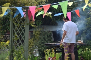 A man in a garden stands over smoky BBQ with bunting in the forefront. Photo Jennifer Ratcliffe 2020